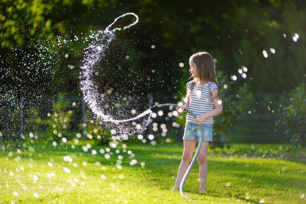 Adorable little girl playing with a garden hose on hot and sunny summer evening